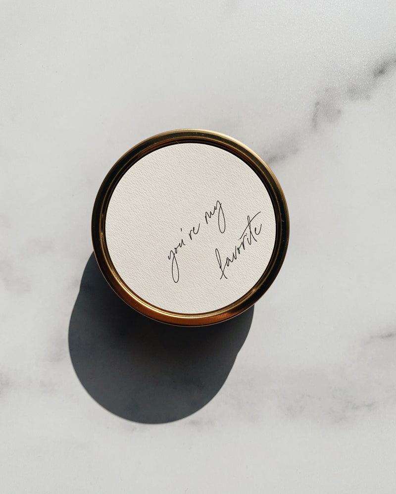 100% soy wax travel candle in gold tin with wooden wicks and you're my favorite on the neutral label in cursive. Harsh shadow on the product image in natural light. 