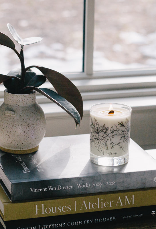 minimalist soy wooden wick candle burning in front of a window on top of books next to a small plant with the sun streaming in behind it
