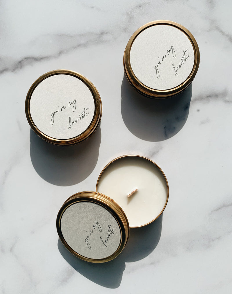 three gold soy wax travel candle tins with wooden wicks and you're my favorite on the neutral label in cursive. Harsh shadows on the product image in natural light. 